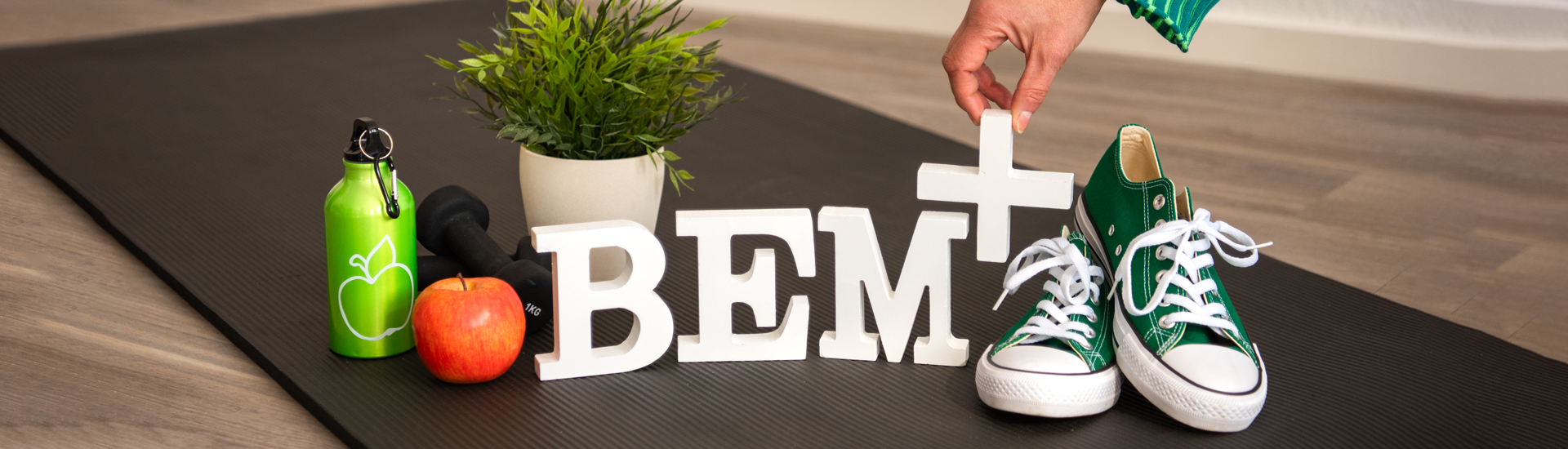 Header BEM+ Die Buchstaben BEM+ in weiß stehend auf einer schwarzen Yoga-Matte auf einem Holzfußboden, rechts daneben grüne Turnschuhe, links daneben ein roter Apfel sowie eine grüne Trinkflasche und schwarze Hanteln, im Hintergrund eine Grünpflanze